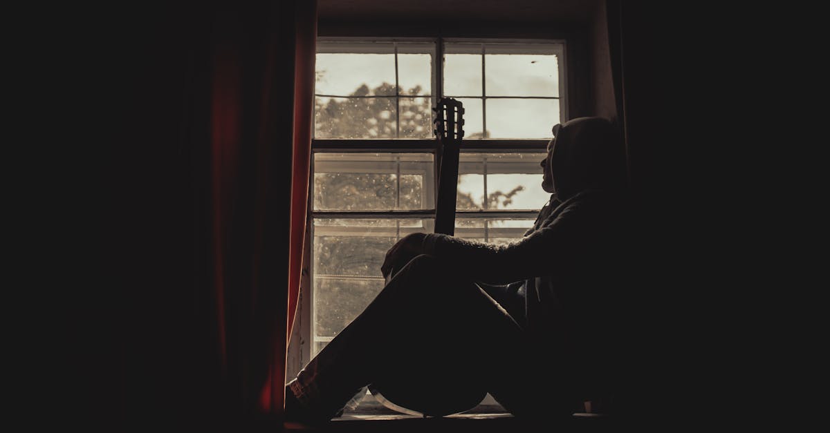 A pensive musician with guitar silhouetted by a window indoors, exuding a moody atmosphere.