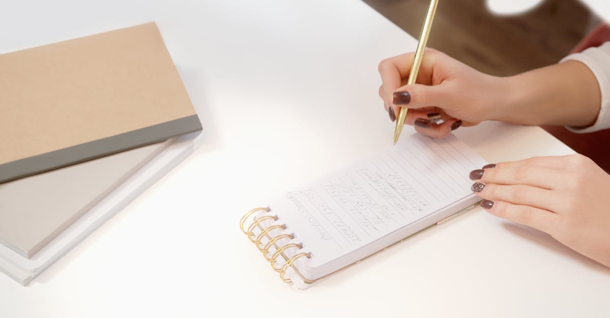A close-up of hands writing in a spiral notebook on a white desk.