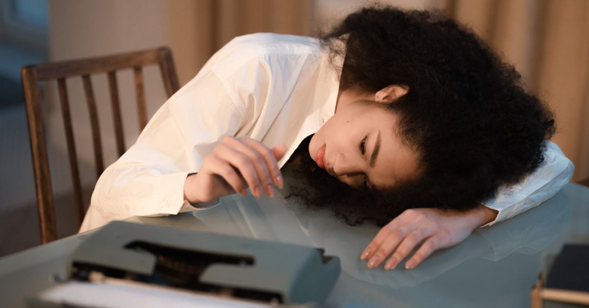 A woman with afro hair in a white shirt rests her head on a table next to a vintage typewriter, appearing fati