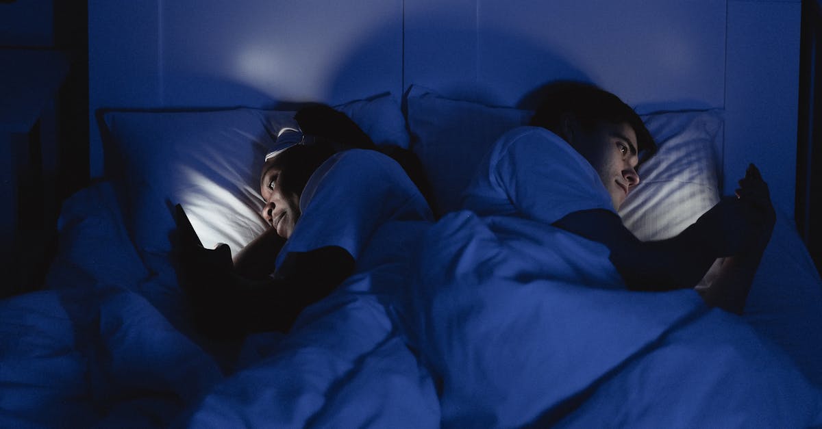 A couple lying in bed using smartphones at night, illuminated by screen light.