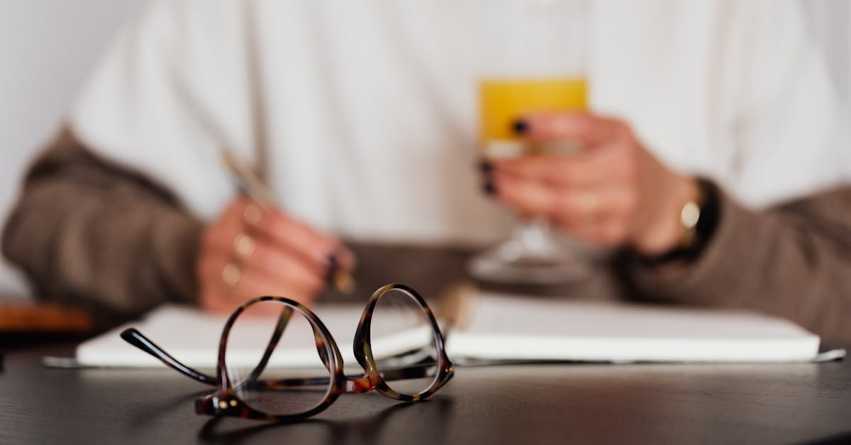 Crop blurred female in stylish sweater with glass of orange juice sitting at table with eyeglasses and writing