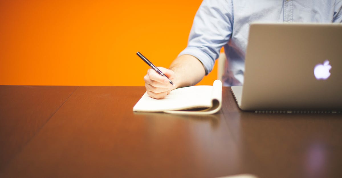 Adult writing notes in a notebook beside a laptop on a wooden desk with orange background.