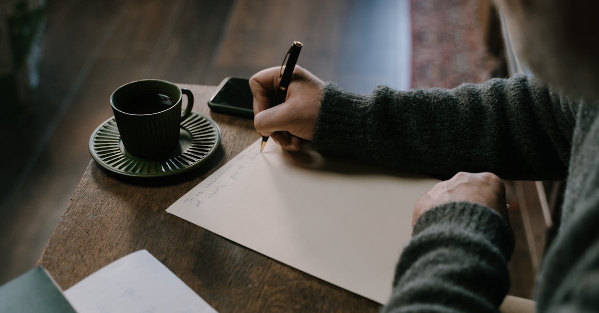 Close-up of a person writing on paper with a cup of coffee on the table.