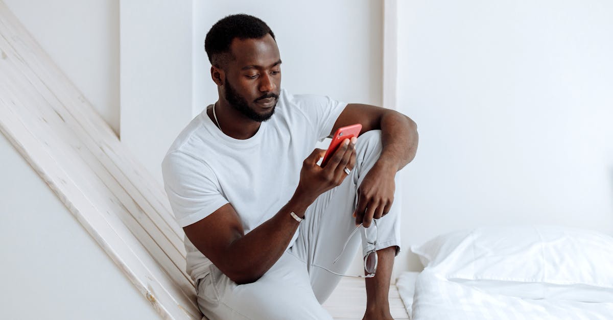 African man sitting on a bed using a smartphone, relaxing indoors.
