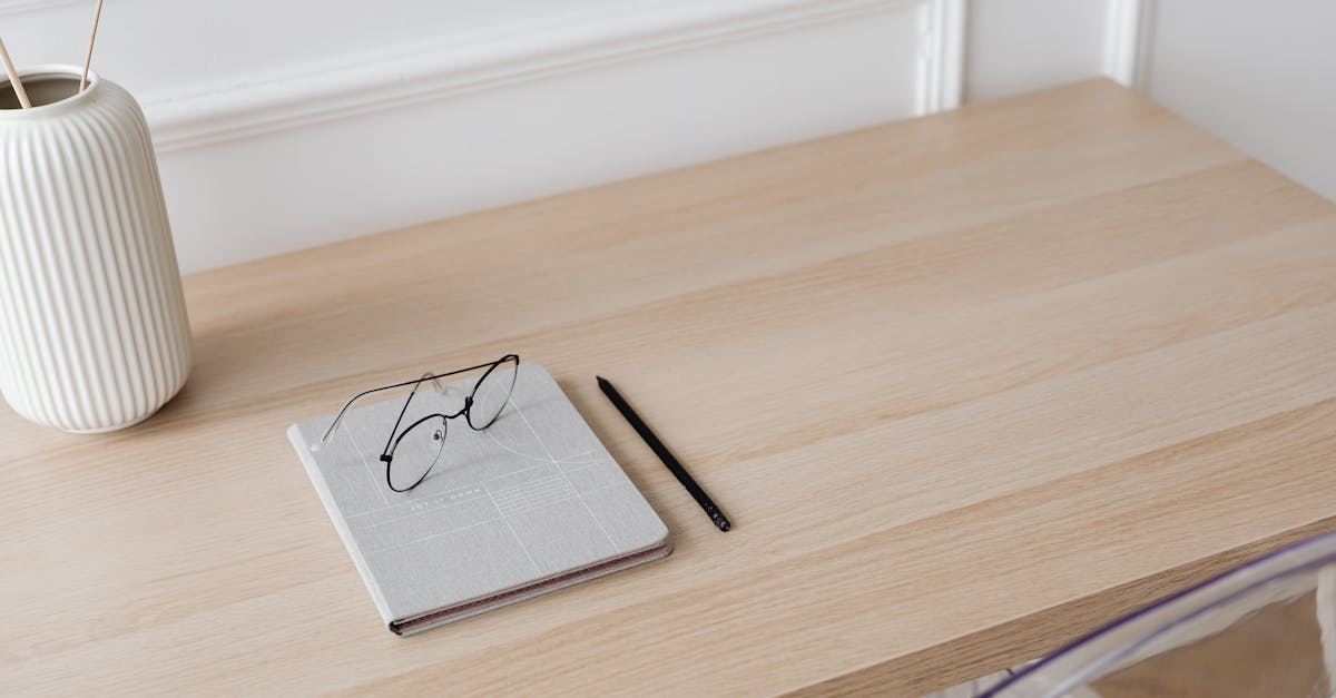 A minimalist workspace featuring a notebook, eyeglasses, pen, and a decorative vase on a wooden desk.