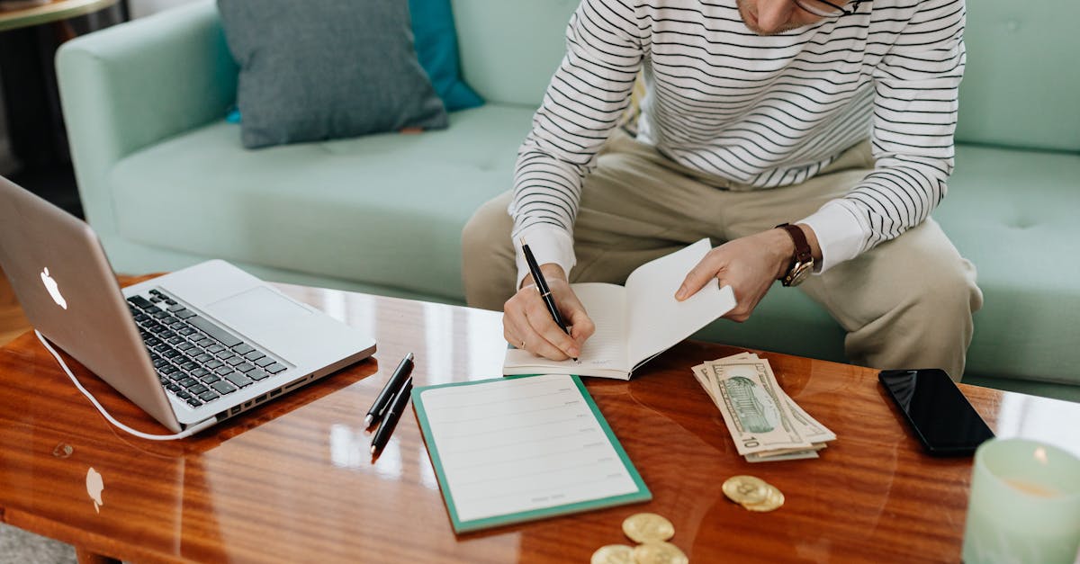 A man in a striped shirt sitting at a wooden table, writing in a notebook with money and a laptop nearby.