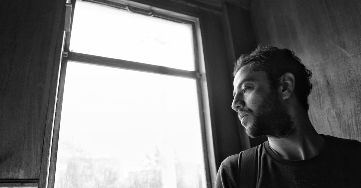 A contemplative man looks out of a window in a black and white photograph, evoking thoughtfulness.