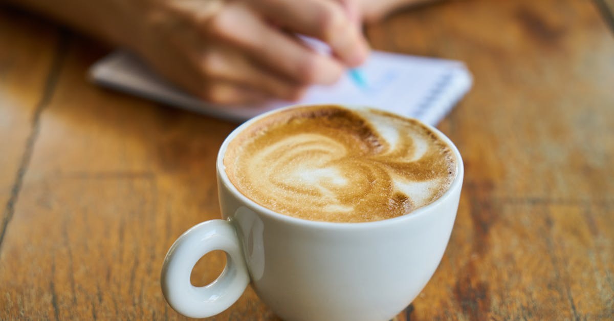 Close-up of a cappuccino cup on a wooden table with someone writing.