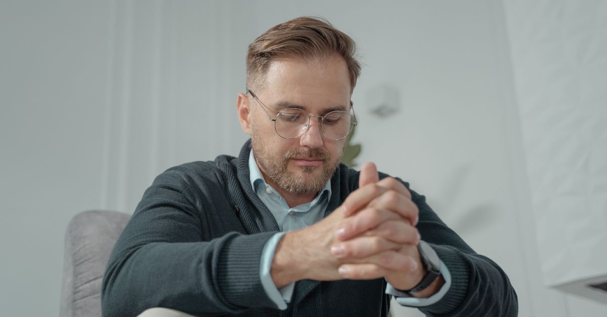 A thoughtful man with glasses and gray sweater sitting indoors, hands clasped.