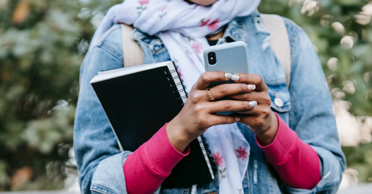 Crop anonymous female in headscarf and jeans jacket with rolled up sleeves and with notebook messaging on cell