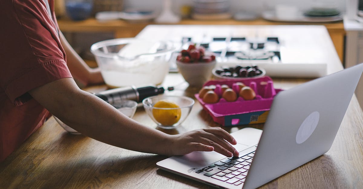 Person using a laptop in the kitchen while preparing baking ingredients. Modern multitasking lifestyle.