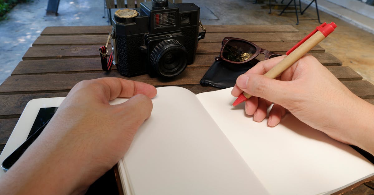 Hands writing in a notebook at an outdoor workspace with a camera and sunglasses.