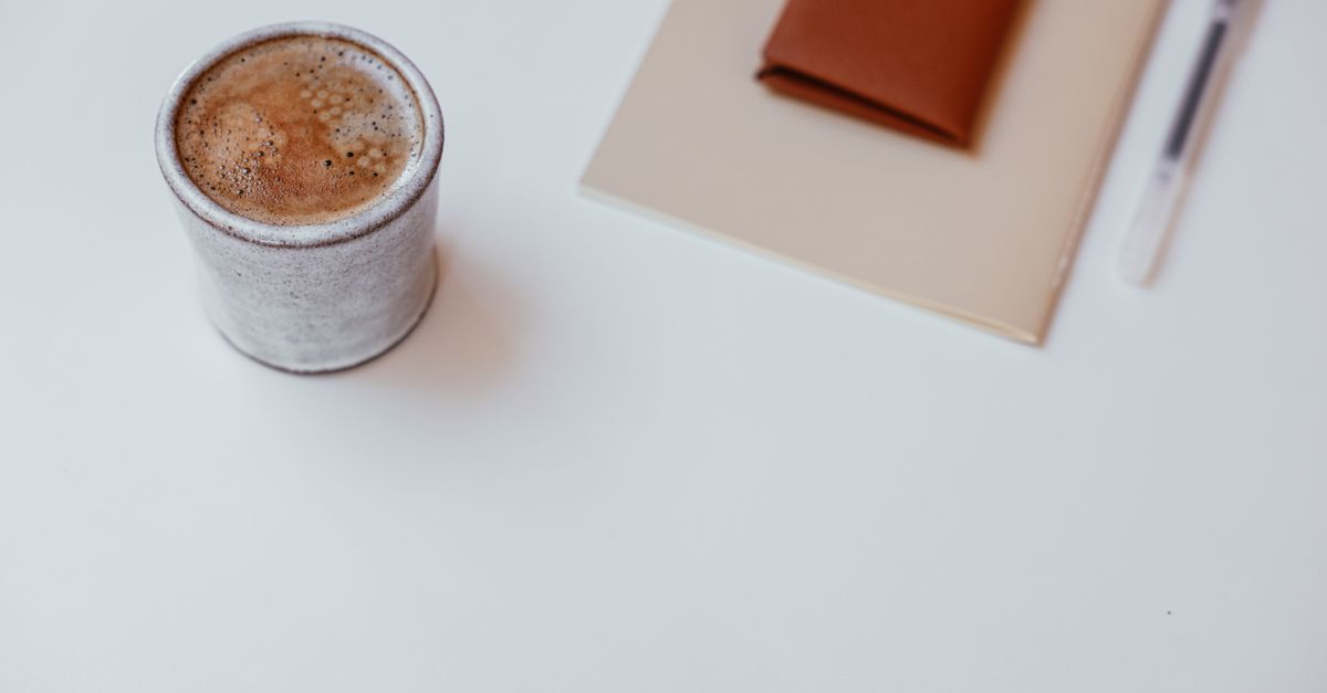 A minimalist setup with a cup of coffee, a notebook, and a pen on a white tabletop.