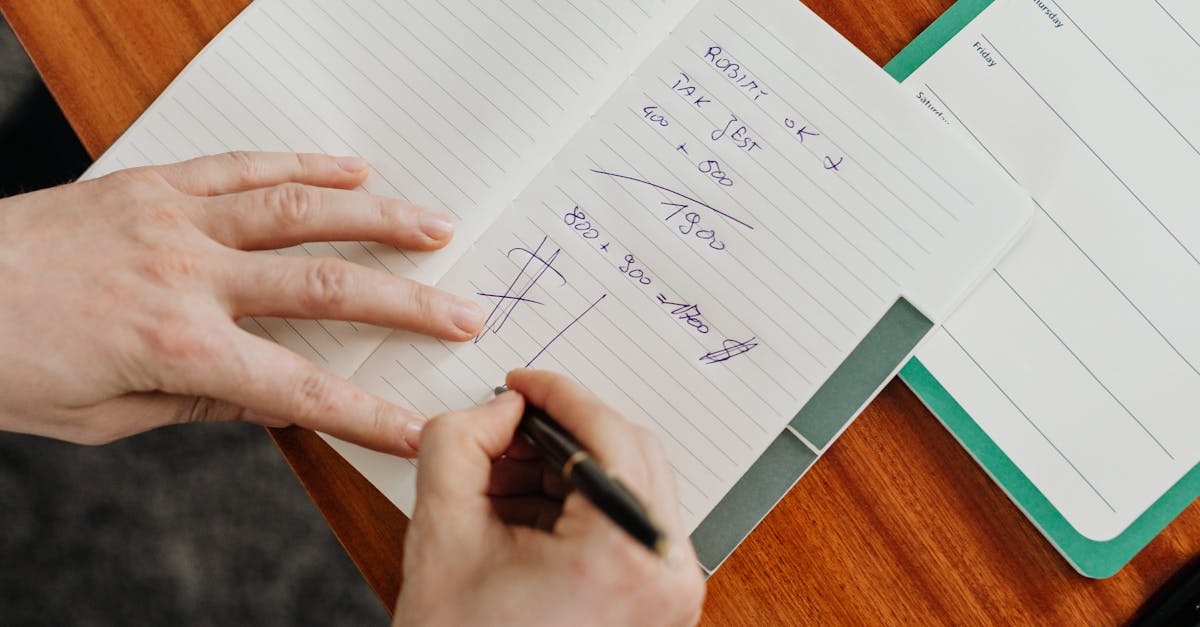 A person writing calculations in a notebook on a wooden desk.