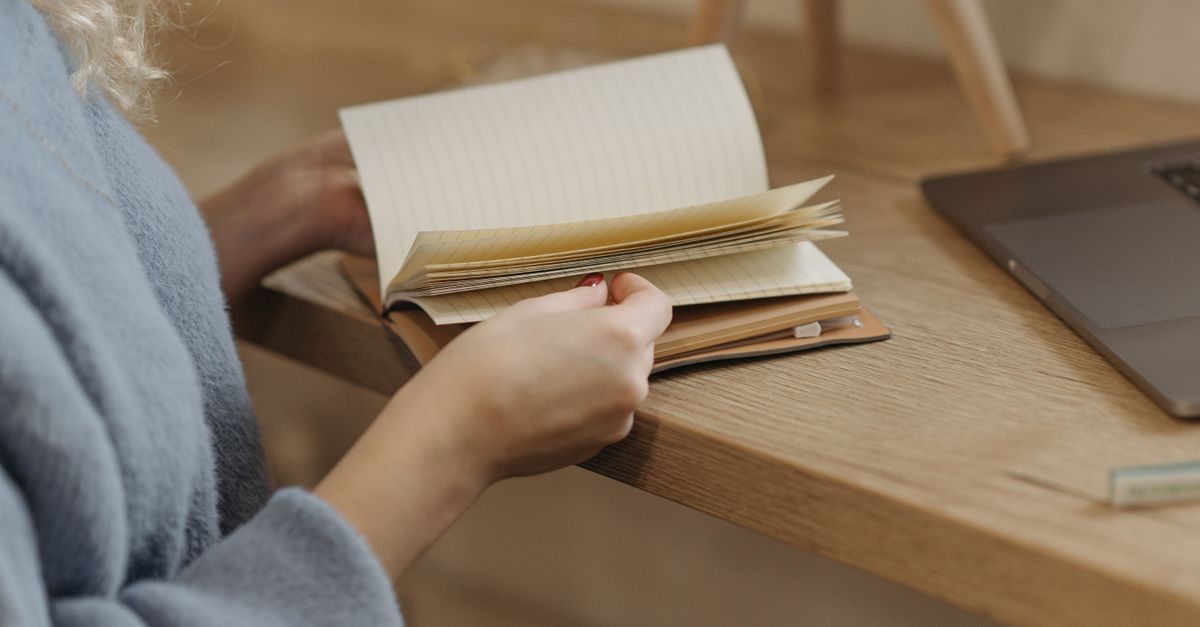 Person flipping through a notebook beside a laptop on a wooden table.