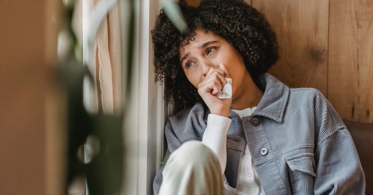 Lonely young black lady with Afro hair looking away while sitting near wooden wall and crying