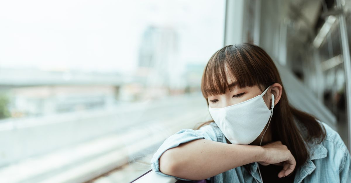 Asian woman wearing a face mask, resting with earphones on a train during the day.