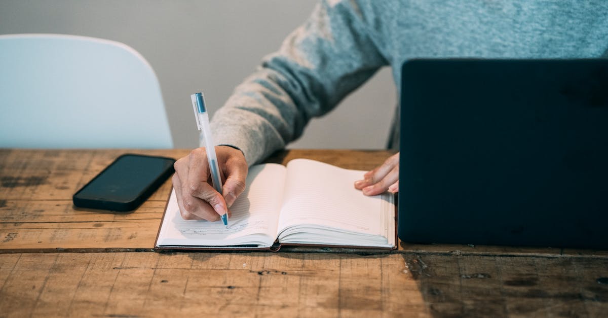 Crop faceless male writing in notebook at wooden table with netbook and blank smartphone on gray background