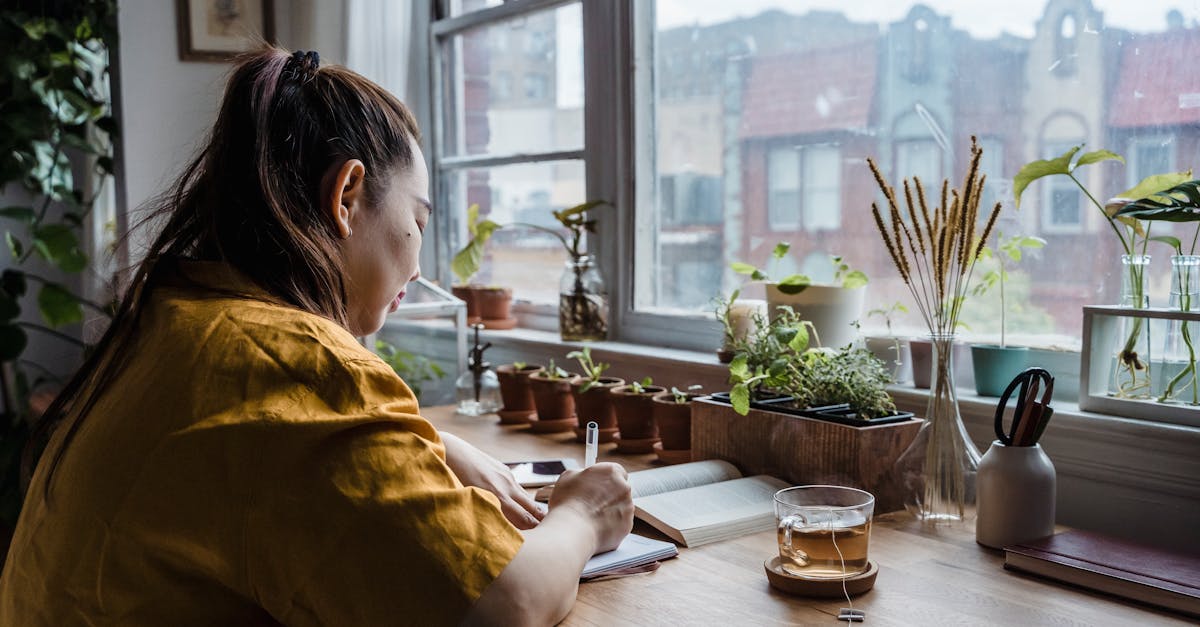 Asian woman writing in notebook in a cozy home office by window with plants.