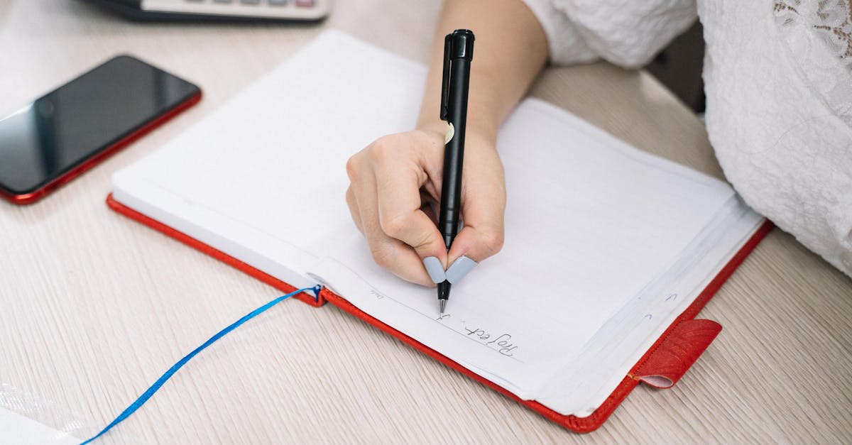 Close-up of an adult hand writing notes in a notebook, with a cellphone and calculator nearby on the desk.