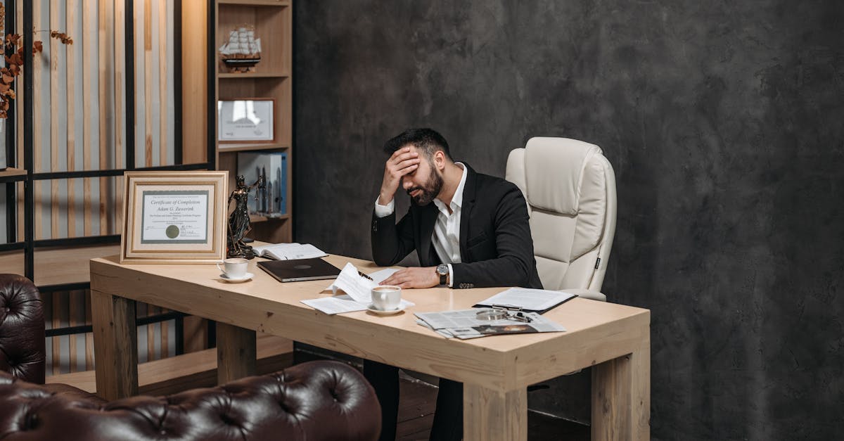 Businessman in a black suit looking stressed at an office desk, holding his head with papers scattered around.